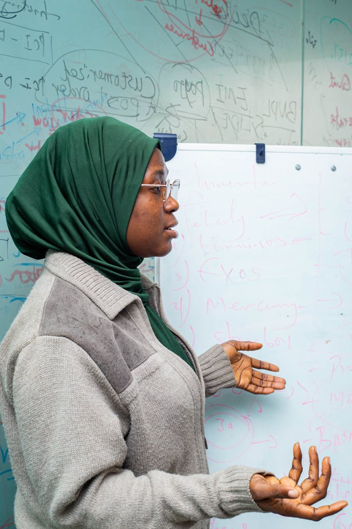 Female teacher in hijab explaining lesson with gestures at whiteboard in a classroom setting.