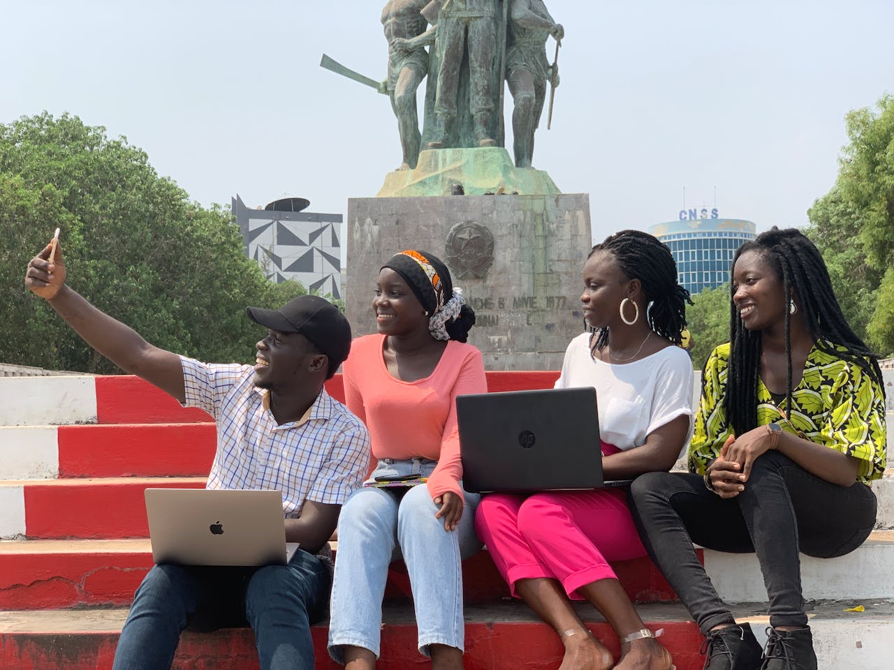 Four friends with laptops smile and take a selfie at a monument in Cotonou, Benin.